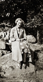 Anna Snygg sitter på en stengärdsgård en solig dag i lång kappa, blommig klänning, knytskor och mössa.