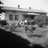 Ernst och Evelina Larsson sitter med dottern Irma och Bertha i Nykvarn (i huckle) i trädgårdsmöbeln framför sitt hus i Utmossen, Fageråkra. I förgrunden står några unga rhododendronbuskar. (Se även EA0388)