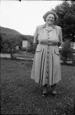 Evelina Larsson i Utmossen, Fageråkra, står i trädgården med syrener och bikupor i bakgrunden. Hon bär en helknäppt klänning med lagda veck och dekorer av vågiga band på krage och ficklock. (Se även EA0393)