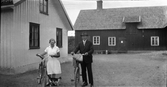 Evelina Larsson på Utmossen, Fageråkra, och Bernt med cyklar invid husgaveln. En katt stryker sig mot Evelinas ben. I bakgrunden står hink och mjölkkanna vid ladugården.