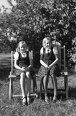 Tvillingsystrarna Elisabeth och Maria Johansson 11 år i likadana kläder. De var fosterhemsplacerade i var sitt hem, men hälsade på varandra. Här är Elisabeth från Eftra på besök hos Maria i Järnmölle, Tvååker. De sitter en solig septemberdag i var sin trädgårdsstol. Elisabeth blev småskollärare i Tvååker under åren 1951-1991 och Maria arbetade som fotvårdsspecialist bland annat på Sannagården.