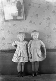 Tvillingarna Maria och Margit (Elisabeth) Johansson fotograferade när de står i soffan i Margits fosterhem. Deras mor födde dem på Lia-hemmet (Fagereds sanatorium), där hon vårdades för TBC, så flickorna hamnade i olika fosterhem. Vid den här tiden bodde Margit i Tvååker hos Augusta Karlsson, som var granne med fotografen, och Maria i Järnmölle i samma socken.