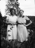 Två unga kvinnor står sommarklädda på en höjd ovanför ett vattendrag. Ingrid Axelsson i Källsmossen till höger har armen om väninnans axel, som möjligen heter Anna Larsson.