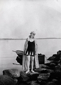 Bad och baddräktsmode.
Ung kvinna iklädd baddräkt, badkappa och badmössa på en stenig strand. 
En roddbåt förtöjd i ett stenblock.