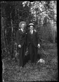 Två välklädda män i hattar, i skogen.
