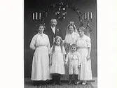 Brudpar. Birger Andersson från Tofta med fru, tärnor och brudnäbbar. I bakgrunden en ärebåge och kandelabrar på pelare.