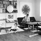 Anna sitter på golvet med dockan medan hennes bror och mamma sitter i soffan i vardagsrummet i Baronbackarna, 1960-tal