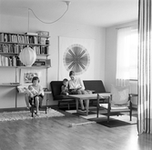 Anna med dockan på trehjuling medan brodern Lars och mamma sitter i soffan i vardagsrummet i Baronbackarna, 1960