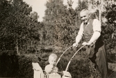 Carl Krantz (1880 - 1956) är på promenad med dotterdottern Eva Pettersson (född 1944, gift Kempe) som sitter i kärran, Stretered år 1945. Carl Krantz var skomakare och undervisade vid Stretereds skolhem.