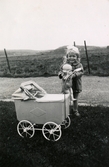 En liten flicka står med sin docka och dockvagn. Namn, årtal och plats är okänt.