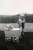 En liten flicka står med sin docka och dockvagn. Namn, årtal och plats är okänt.