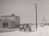 Tvååkers Elaffär AB och ett bostadshus i funkisstil vid Fastarpsvägen i Tvååker vintertid. I förgrunden en hästdragen vagn lastad med (sädes)säckar framför sadelmakare Georg Olssons tomt, Fastarpsvägen 24. Ca 1940.