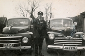 Fritiof (elev boende på Stretereds skolhem) iförd polisuniform och skärmmössa, står mellan två taxibilar vid Kållered station 1940-tal. Han fick 