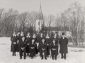 Konfirmandgrupp ute i snön med Skällinge kyrka i bakgrunden. Prästen bär hög hatt.