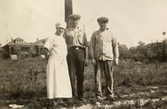 Tre personer står på en åker, troligtvis i Alaska 1924. En byggnad ses i bakgrunden.