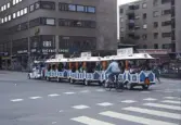 Turisttåg kör i sydvästra hörnet av korsningen Drottninggatan-Rudbecksgatan, 1987