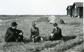 Gunnilbo sn, Bussbyn.
Tre män sitter i gräset framför lador. 1930-tal.