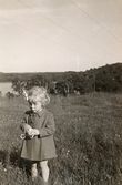 Eva Pettersson (född 1944, gift Kempe) står på en sommaräng, finklädd i kappa och med en blomma i håret år 1946. I bakgrunden ses Tulebosjön. Dotter till Bror och Rosa Pettersson.