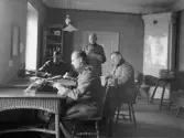 Fyra uniformerade män, militärer, utför administrativa uppgifter vid bord i rum med kakelugn. Närmast en man som skriver på skrivmaskin, en sitter och slår i en pärm, en annan antecknar och den fjärde står med kuvert eller andra handlingar i händerna. Uppgifter om bilden saknas.