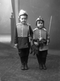 Fotografens två söner Åke t h och Bo t v utklädda i uniformer med kaskar och värjor. Åke håller i en docka.