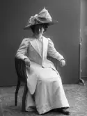 Fotografens hustru modist Elin Björkström sittande i ljus dräkt med bredbrättad stråhatt med enorm rosett.
