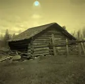 Ekonomibyggnad med sadeltak av rundtimmer, gärdgård. och åkermark.