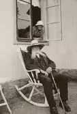 Anna Larsson tittar ut genom fönstret på sin make Axel Larsson i gungstolen på Pettersbergs gård, 1938