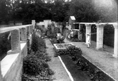 Trädgården till Gerlachska villan med pelarrader, rabatter och en damm i mitten. Bilden tagen från väster. Flickan som står till höger är troligen Margit Gerlach och kvinnan bakom henne som sitter på en bänk är troligen Anna Bagge.