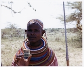Läkerol på segertåg genom Afrika.
Afrikansk kvinna håller en Läkerol orginal ask.
Kvinna har färgrika smycken runt halsen, stora örhängen samt håller ett spjut.