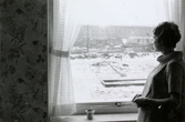 Ninnie Tobiasson (född Johansson), gravid med dottern Ilse, står och tittar ut från sitt lägenhetsfönster på Bågskyttegatan 3g i Mölndal 1959/1960. Utanför fönstret ses bostadsområdet (Bågskyttegatans jämna nummer) som är i sluttampen av nybyggnation.