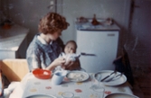 Ninnie Tobiasson (född Johansson, 1937) testar att lära dottern Ilse (född 1960, gift Glimberg) att äta mat, Krokslätt 1960. Matningen gick sådär och Ilse sög mest in maten. Troligtvis hemma hos mormor och morfar som bodde vid nedförsbacken mitt emot Krokslättsskolan.