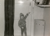 3-åriga Ilse Tobiasson (född 1960, gift Glimberg) står i hallen och är på väg ut för att leka, Krokslättsgatan 3 år 1963. Hon är klädd i hemmastickad matchande tröja, mössa och halsduk. Till höger ses en tvättkorg som av utrymmesskäl fick stå i hallen. Lägenheten var en omodern 1:a på bottenvåningen.