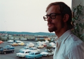 Esbjörn Hansson fotas i profil ståendes på balkongen, Havrekornsgatan i Bifrost cirka 1972-1974. I bakgrunden ses en parkering.
