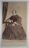 Visitkortsporträtt av Christina Zachau. Fotografiet sitter i fotoalbum som tillhört Gertrud Zachau. 
Kvinnan avbildas sittandes på stol med höger arm vilande på ett bord.