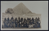 Gruppbild av turister i två rader som poserar framför en pyramid. Turisterna sitter på kameler. Den främre radens kameler hukar, den bakre stående. Möjligen är ledsagare stående bredvid.

Text kortets baksida 