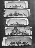 Dörröverstycken som ingår i en svit väggfält och delar av väggfält, 14 stycken, målade med limfärg på grovt papper och uppklistrade på grov, tät linneväv. Målade i en något naivistisk stil med fina detaljer. Samtliga delar med målad, profilerad ram i bronsimiterande ockratoner Från  Freluga i Hälsingland. Målningarna är utförda av Jonas Hartman ca. 1775-1804. Dörröverstycken från 1799.