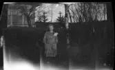 Flicka, troligtvis Karin Sundling vid häck framför hus.
