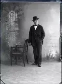 Ateljébild av Karl Johansson i helfigur iklädd kostym, fluga och hatt vid en stol. Beställare:  F Hjalmarsson, Falkenberg.