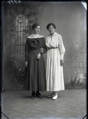 Ateljébild med två stående kvinnor i klänningar som krokat arm. Beställare: Fröken Palmér, Stafsjö.