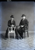 Ateljébild, Två sittande unga män i hatt och kostym. Den ene sitter på en taburett med armarna över dess ena gavel. Beställare och troligen avbildad: H Larsson. Har även beställt bildnr GB2_1774.