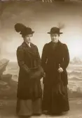 Två damer stående i helfigur. Ateljébild. Den ena har pälskappa och muff samt hatt med plym. Den andra damen bär kappa och hatt med fjäder, samt håller ett paraply i vänster hand. Beställare: Augusta Jansson. Lahall. (Se även bildnr GB2_5619)