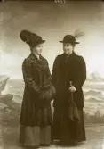 Två damer stående i helfigur. Ateljéfoto. Den ena har pälskappa och muff samt hatt med plym. Den andra damen bär kappa och hatt med fjäder, samt håller ett paraply i vänster hand. Beställare: Augusta Jansson. Lahall. (Se även bildnr GB2_5618)