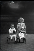Ateljébild.  En liten pojke och äldre flicka i helfigur. Pojken sitter på en pall med leksak i handen. Flickan står bakom en pall och håller en stor docka, som sitter på pallen framför henne. Beställare: Frida Samuelsson. (Salomonsson )