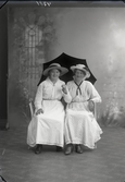 Ateljébild. Två kvinnor iförda hattar och ljusa, småmönstrade klänningar sitter och håller ett svart paraply bakom sig. Beställare: Frida Salomonsson, Frk. Petterssons möbelaffär, Falkenberg.