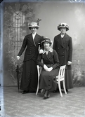 Ateljébild. Tre kvinnor i helfigur där hon i mitten sitter på en taburett. Samtliga bär smalbrättade, rikt dekorerade hattar och ytterkläder med stora knappar. Den ena kvinnan stödjer sig på ett paraply. Beställare: Ida Andersson, Gunnarp.
