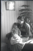 Mor med liten baby sitter i en soffa i hemmiljö med en krukväxt bakom dem. Beställare: Handl. Georg Jönsson Falkenberg.