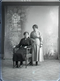 Ateljébild. Två kvinnor i helfigur varav en sitter. Beställare: Nanny Nylander, Falkenberg. ( Rika Skoog)