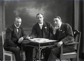 Ateljébild. Tre män i halvfigur som sitter kring ett bord där en tidning ligger. Två bär blommor på kavajslagen. Beställare och sannolikt avbildad: Robert Eriksson, Falkenberg, Valskvarnen. (Se även bildnr GB2_5919)