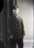 Ateljébild. Lotsförman Adolf Ekman, Falkenberg, stående i helfigur iklädd lotsuniform med skärmmössa.