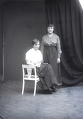 Ateljébild. Två unga kvinnor i helfigur varav en sitter på en taburett. Beställare: Lucia Larsson, Sanddynerna 10, Falkenberg.